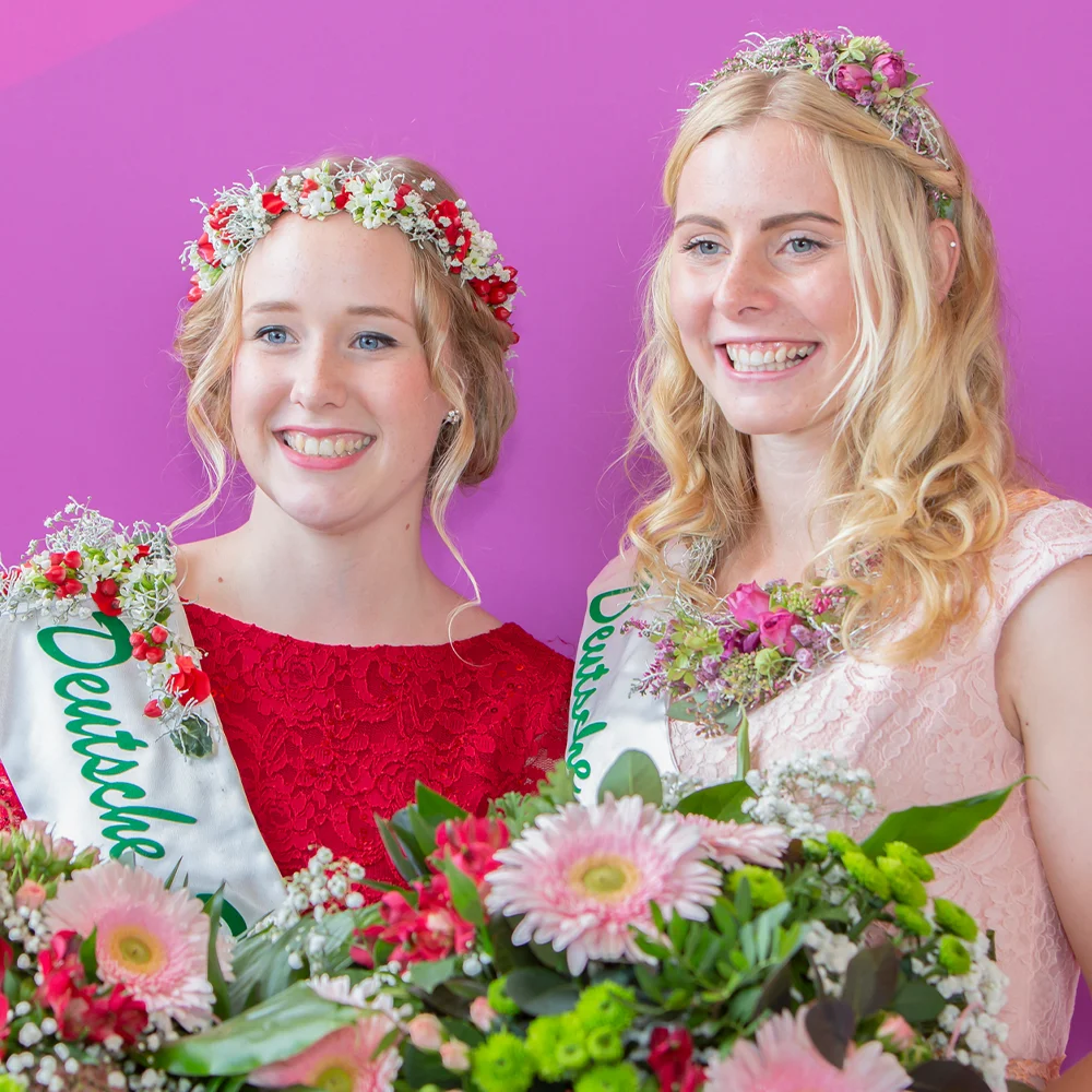 Eventfotografie von zwei jungen Frauen, die als Blumenprinzessinnen gekrönt sind, mit Blumenkronen und Sträußen, aufgenommen vor einem leuchtend violetten Hintergrund.