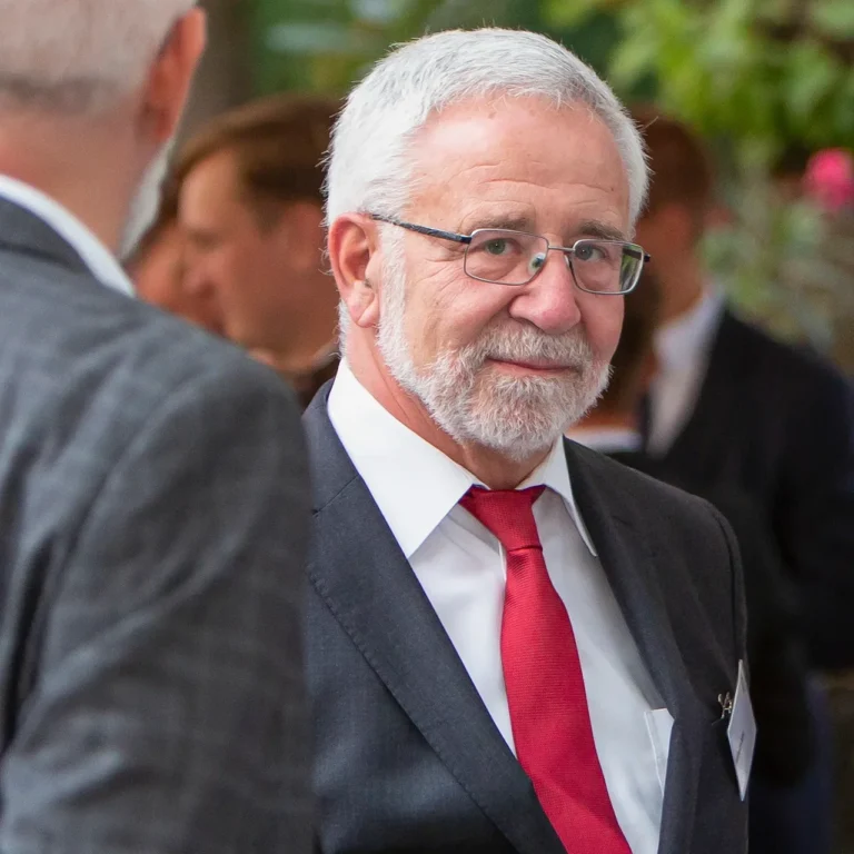 Ein Eventfoto eines älteren Mannes in einem dunklen Anzug mit roter Krawatte und Brille, der bei einer Veranstaltung im Gespräch ist.