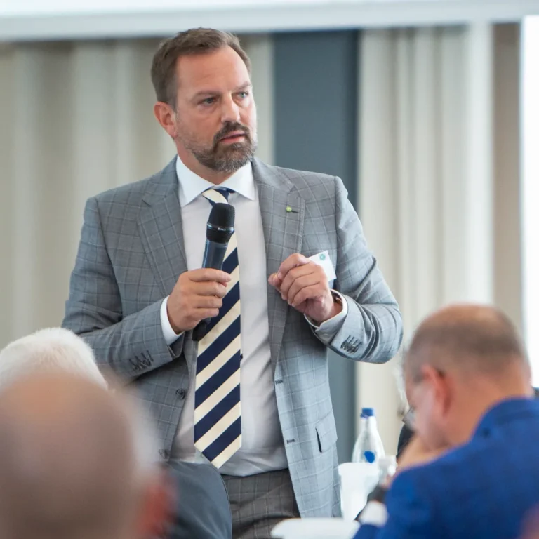 Ein Eventfoto eines Mannes in einem grauen Anzug mit gestreifter Krawatte, der bei einer Konferenz spricht und ein Mikrofon hält.