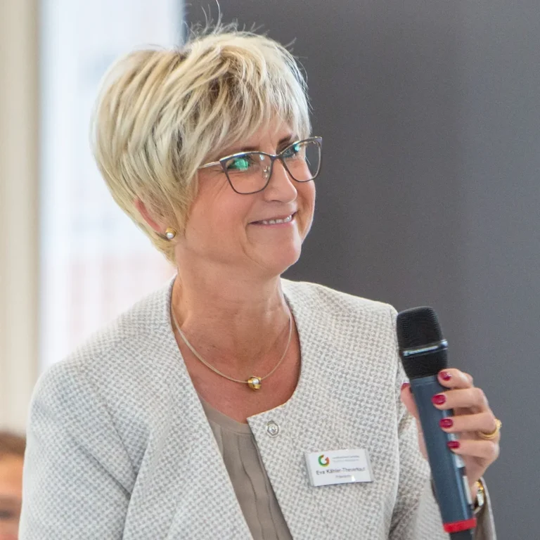 Ein Eventfoto einer Frau in einem hellen Blazer, die bei einer Konferenz spricht und ein Mikrofon hält.