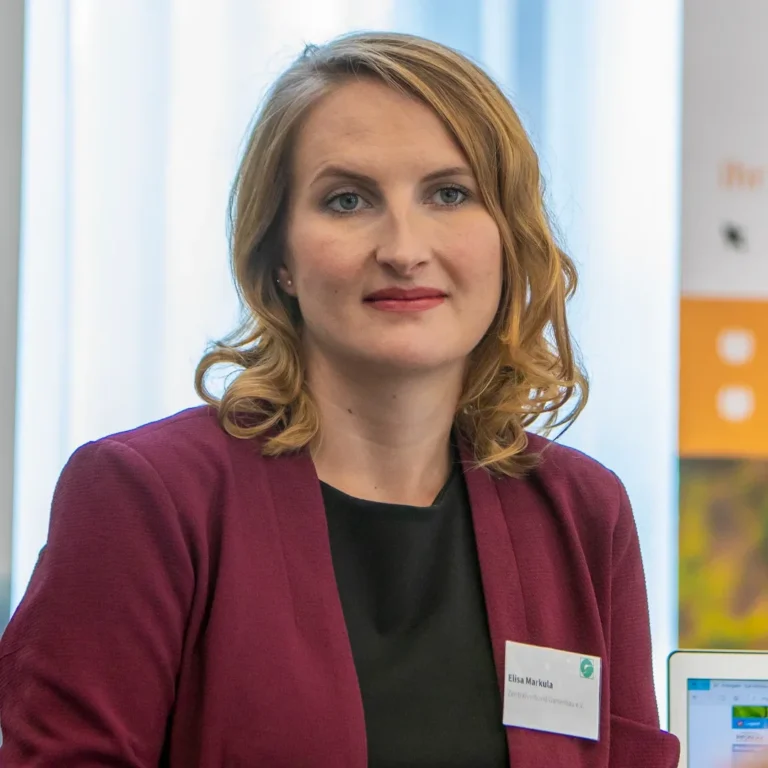 Ein Eventfoto einer Frau in einem weinroten Blazer, die bei einer beruflichen Veranstaltung am Tisch sitzt und aufmerksam zuhört.
