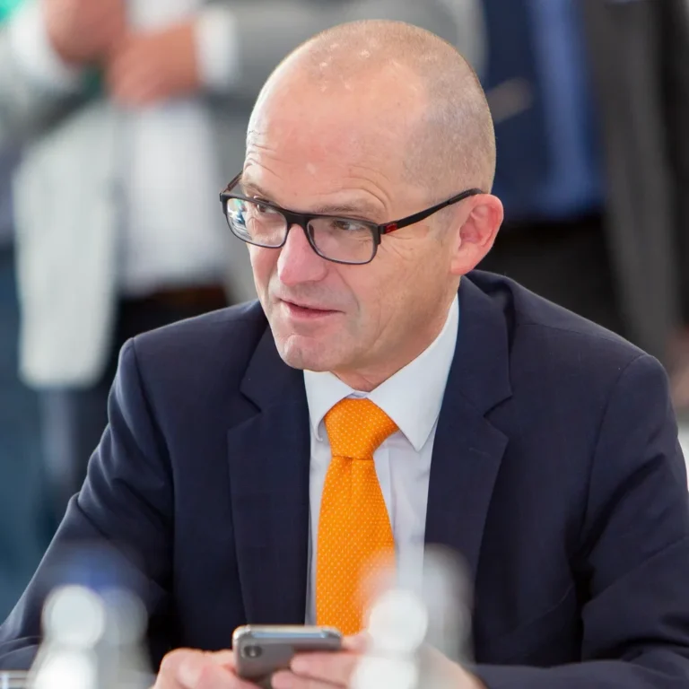 Ein Eventfoto eines Mannes in einem dunklen Anzug mit orangefarbener Krawatte und Brille, der bei einer Konferenz sitzt und auf sein Handy schaut.