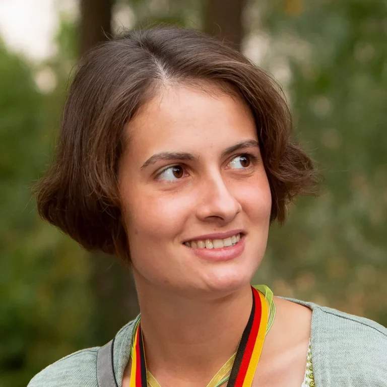 Ein Eventfoto einer jungen Frau in traditioneller Tracht mit einer Medaille um den Hals, die auf einem Fest im Freien lächelt.