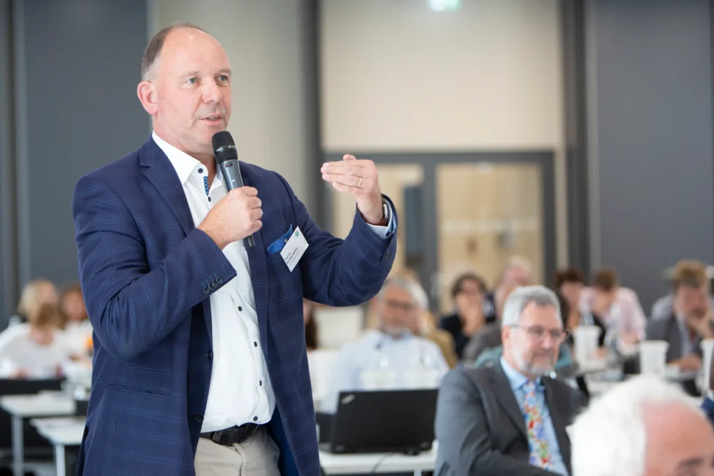 Ein Eventfoto eines Mannes in einem blauen Anzug, der eine Rede mit einem Mikrofon bei einer Konferenz hält.