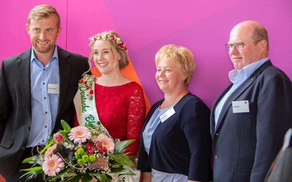 Ein Eventfoto von vier Personen, darunter eine junge Frau in einem roten Kleid mit einer Blumenschärpe, die als Deutsche Blumenfee gekrönt ist, posierend vor einem violetten Hintergrund.